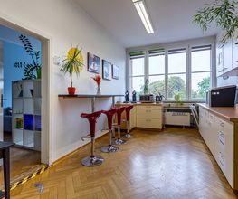 Küche mit Sitzmöglichkeiten. Mikrowelle, Wasserkocher und Kaffeemaschine stehen in der Küche. Mehrere Pflanzen stehen verteilt in der Küche. An der Wand befinden sich Kaffee-Bilder.