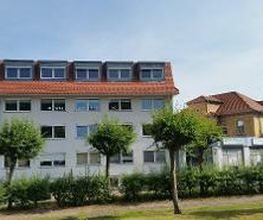 Weißes Gebäude hinter Grünfläche und Bäumen.