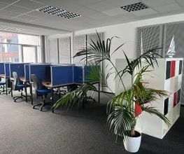 Ein heller Raum mit Arbeitsplätzen, die durch blaue Trennwände voneinander getrennt sind. Vor jedem Arbeitsplatz steht ein Bürostuhl. Rechts stehen ein Regal mit bunten Fächern und eine große Zimmerpflanze in einem weißen Topf.