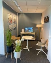 Beratungsbereich mit Tisch und 2 Stühlen. Im Hintergrund ein Regal mit TV, links stehen 2 Pflanzen, an der Wand hängen 2 Plakate.