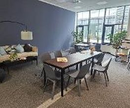 Gemütlicher Aufenthaltsraum mit Tischen und Stühlen, sowie einem modernen Sofa, einem Sitzkissen, einer stilvollen Stehlampe und mehreren Zimmerpflanzen.