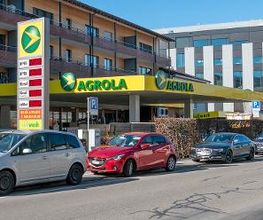 AGROLA Tankstelle in Zug; auf linker Seite Tankstelle hinter geparkten Autos; grosses Totem mit AGROLA Logo und digitalen Preisschildern sowie Selfwash Logo