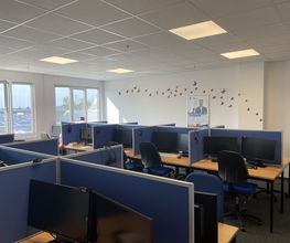 Schulungsraum mit mehreren Arbeitsbereichen, die durch blaue Trennwände voneinander abgegrenzt sind und mit 2 PC-Bildschirmen, Tastatur, Maus und Headset ausgestattet sind. Fensterreihe und gebastelte Schmetterlinge an der Wand.