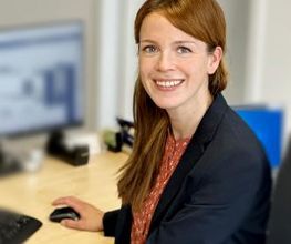 Portrait von Mandy Altmann, Teamassistentin am WBS Standort Güstrow. Sie hat lange, hellbraune Haare und sitzt am Schreibtisch vor ihrem Computer und lacht fröhlich in die Kamera.
