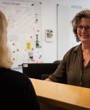 Eine Frau mit lockigem Haar und Brille steht lächelnd hinter einem Empfangstresen. Ihr gegenüber ist der Rücken einer anderen Person zu sehen. Im Hintergrund hängen ein Jahresplaner und eine Pinnwand an der Wand. Der Raum wirkt hell und freundlich.