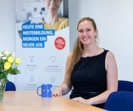 Eine Frau mit blonden Haaren und dunkelblauem Top sitzt an einem Tisch. Vor ihr stehen eine blaue Kaffeetasse und ein Strauß gelber und weißter Blumen.