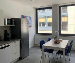 Eine Küche mit Mikrowelle, Kaffeemaschine, Kühlschrank und einem weißen Tisch mit 4 Stühlen, auf dem eine Blume und eine Obstschale steht.