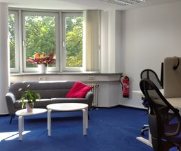 Arbeitsraum mit höhenverstellbaren Tischen, mit jeweils 2 Bildschirmen, einem Bürostuhl, Tastatur, Maus und Headset. Daneben ein Sofa mit Beistelltischen und Pflanzen.