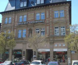 gelbes Haus mit dem Namen Haus des Handwerks. Im unteren Bereich des Hauses sind Schaufenster und davor stehen Autos. Im oberen Bereich sind mehrere Fenster und der Schriftzug Haus des Handwerks.