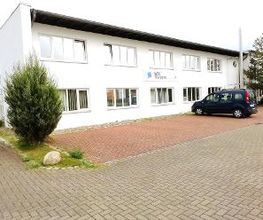 Zweistöckiges Schulungsgebäude WBS TRAINING in Waren. Davor ist ein Parkplatz mit einem geparkten AUto zu sehen.