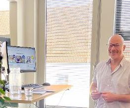 Ein Mann mit Brille und hellem Hemd steht lächelnd vor einem Stehpult. Auf dem Pult sind ein Monitor, eine Glaskaraffe, ein Glas und Unterlagen. Im Hintergrund sind Fenster mit Jalousien und das Dach eines Gebäudes zu sehen. Links steht eine Zimmerpflanze