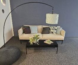 Gemütliches, helles Sofa mit Kissen. Davor steht ein schwarzer tiefer Tisch mit Blumen und Zeischriften. stilvolle Stehlampe und graues Sitzkissen.