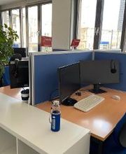 Arbeitsplätze mit blauen Trennwänden, 2 Bildschirmen, Tastatur, Maus und Headset. Der Raum hat viele Fenster. Es befindet sich ein weißes Regal auf dem eine grüne Topfpflanze und eine Getränkeflasche steht in dem Raum.