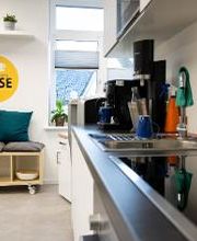 Voll ausgestattete Küchenzeile. Kleine Sitzbank mit Kissen, darüber hängt ein gelber, runder Aufkleber mit der Aufschrift Mach einfach mal Pause und einer weißen Kaffeetasse.