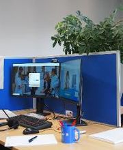 PC-Arbeitsplatz mit blauer Trennwand sowie 2 Bildschirmen, einer Tastatur und einem Headset. Auf dem Schreibtisch befinden sich ein Notizbuch, Papier uns Stifte sowie eine blaue Tasse.