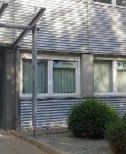 Ein modernes Gebäude mit Metallfassade. Links befindet sich ein Eingang mit blauem Türrahmen und Glasüberdachung. Rechts steht ein Schild mit Logos und „WBS Training“. Vor den Fenstern sind Büsche und ein Kiesbett angelegt. Der Bereich wirkt gepflegt.