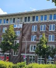 Außenansicht des Gebäudes. Brauner Klinker und weiße Fensterrahmen. WBS TRAINING Schild an der Fassade. Grüne Bäume und Sträucher.