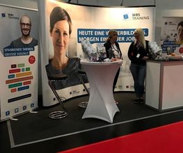 WBS-TRAINING Messestand mit Plakataufstellern und 2 Beraterinnen an einem Stehtisch.