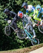 Mehrere BMX-Fahrer springen gleichzeitig über eine Rennstrecke während eines Wettbewerbs. Die Szene vermittelt Geschwindigkeit und intensive Konkurrenz.