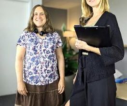 2 Frauen stehen neben einem Tisch. Frau rechts hält eine Klemmmappe. Im Hintergrund steht eine Zimmerpflanze und eine Stehlampe.