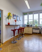 Küche mit Sitzmöglichkeiten. Mikrowelle, Wasserkocher und Kaffeemaschine stehen in der Küche. Mehrere Pflanzen stehen verteilt in der Küche. An der Wand befinden sich Kaffee-Bilder.