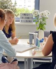 Hier sitzen drei Damen an einem Tisch in einem hellen, gemütlichen Raum und unterhalten sich.