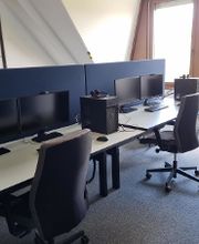 Ein Schulungsraum mit mehreren Arbeitsbereichen, die durch blaue Trennwände voneinander getrennt sind. Jeder Arbeitsplatz besteht aus 2 PC-Bildschirmen, Tastatur und Maus sowie Headset und blauen Trennwänden.