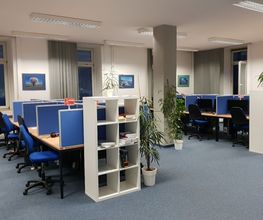 großer Raum mit mindestens 9 Arbeitsplätzen. Alle jeweils mit 2 Bildschirmen, einem Bürostuhl und Tastatur. Im Hintergrund sind mehrere Fenster, im Vordergrund stehen Regale. Mehrere Zimmerpflanzen sind im Raum verteilt. Blauer Teppichboden.