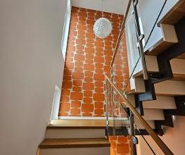 Moderne Holztreppe mit Glasgeländer vor einer kontrastreichen, orange-gemusterten Design-Tapete und einer kugelförmigen Design-Hängeleuchte