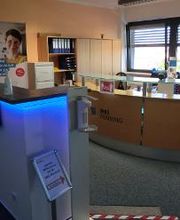 Empfangsbereich mit 2 Theken, einer mit LEDs und Händedesinfektion, die andere aus Holt mit Glasplatte. Hinter der Holztheke sind 2 Arbeitsplätze und mehrere Holzschränke