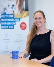 Eine Frau mit blonden Haaren und dunkelblauem Top sitzt an einem Tisch. Vor ihr stehen eine blaue Kaffeetasse und ein Strauß gelber und weißter Blumen.