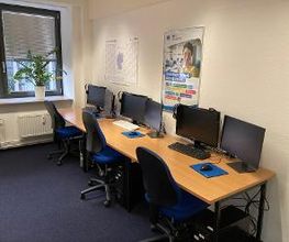 3 Arbeitsplätze mit jeweils 2 Bildschirmen, einem Bürostuhl, Tastatur, Maus und Headset. An der Wand ein Kalender und ein WBS TRAINING Plakat.