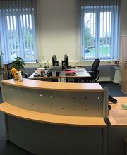 Empfang und Arbeitsplatz der Mitarbeitende. 2 Schreibtische mit Bildschirmen, Pinnwände an der Wand, Sitzecke, Schrank und Fenster mit Sonnenschutz.