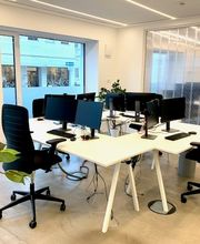 Hell und offen gestalteter Schulungsraum mit weißen, modernen Schreibtischen und schwarzen, modernen Schreibtischstühlen. Monitore auf den Tischen. Helle Glasfront sorgt für viel Licht. Heller Holzfußboden. Grünpflanze.