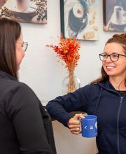 2 Frauen sitzen sich gegenüber. Die rechte Frau hält eine blaue Tasse in der Hand und trägt eine blaue WBS GRUPPE Strickjacke. Sie Lächelt die andere Frau an. Im Hintergrund steht eine Vase mit Trockenblumen. An der Wand hängen Kaffeebilder.