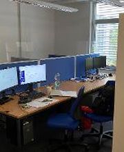 Mehrere Arbeitsplätze mit jeweils 2 Bildschirmen, einem Bürostuhl und Tastatur und Maus. Ein Regal dient als Raumtrenner. An der Wand hängt ein Plakat der WBS TRAINING.