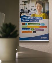 Eingangsbereich mit Poster an der Wand auf dem die Weiterbildungsthemen zu sehen sind. Davor steht ein Regal mit einer kleinen Topfpflanze und einer Lichterkette.