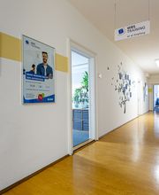 Flur mit mehreren Plakaten an den Wänden und Türen die in verschiedene Zimmer führen. Am Ende des Flures steht ein Roll up Banner und an der Decke hängt ein Schild mit Wegpfeil zum Empfang.