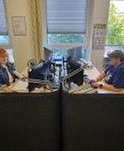 Empfangsbereich mit 2 Mitarbeitenden, die an ihren Schreibtischen vorm Computerbildschirm sitzen und gut gelaunt arbeiten. Geöffnetes Fenster mit Blick ins Grüne.