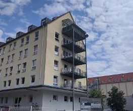 gelbes Gebäude mit vielen Fenstern und Balkonen an der Seite. Unten steht eine Tafel mit Beschilderungen und ein paar Autos.
