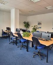 Ein Schulungsraum mit mehreren Arbeitsbereichen. Jeder Arbeitsplatz besteht aus 2 PC-Bildschirmen, Tastatur und Maus sowie Headset und einem blauen Bürostuhl.
