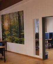 Ein PC-Arbeitsplatz, ein Bild einer Waldszene und eine Tür in einen anderen Raum, in dem sich ebenfalls PC-Arbeitsplätze befinden.