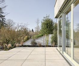 Großzügige Terrasse mit direktem Übergang zur Natur: Die klare Geometrie der Bodenplatten trifft auf weiche Landschaftsformen und üppige Begrünung. Glasfassade und Pflanzung verschmelzen Innen- und Außenraum.