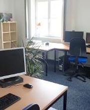 Schulungsraum mit 4 Arbeitsplätzen jeweils mit 2 Bildschirmen, einem Bürostuhl, Tastatur und Maus. An den Wänden hängen Plakate. In der Ecke steht ein Regal mit einer Pflanze.