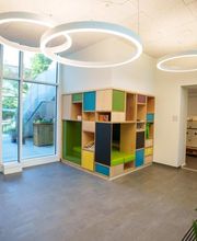 Helles Foyer im Fröbel-Kindergarten Essener Straße mit bodentiefen Fenstern, runden Deckenleuchten und farbigem Holzregal mit Sitznischen.
Fröbel-Kindergarten Essener Straße, Hamburg, © Fröbel, Foto: Martin Arnold. Alle Rechte vorbehalten.