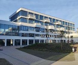 Weißes Gebäude mit mehreren Fenstern und Balkonen. Davor Grünflächen und Pflastersteine