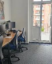 Schulungsraum mit 4 Arbeitsplätzen mit jeweils 2 Bildschirmen, einem Bürostuhl, Tastatur, Maus und Headset. An der Wand ein Kalender und ein Plakat.