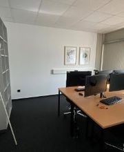 Schreibtische im Trainingsraum mit jeweils 2 PC-Bildschirmen, Tastatur, Headset, Maus und zwei Bildschirmen, Garderobe steht unmittelbar neben einem Bücherregal.