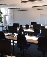 Schulungsraum mit mehreren Tischreihen an den blaue Bürostühle stehen. Auf den Tischen befinden sich Computerbildschirme sowie Tastatur und Maus.