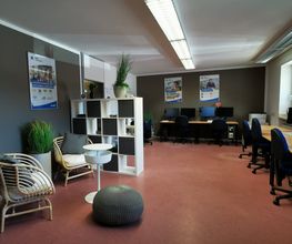 Schulungsraum mit mehreren Arbeutsplätzen mit jeweils 2 Bildschirmen, einem Bürostuhl, Tastatur und Maus. Links stehen 2 Sessel und ein Beistelltisch. An der Wand hängen 3 Plakate. Ein Regal trennt den Sitzbereich von der offenen Tür.